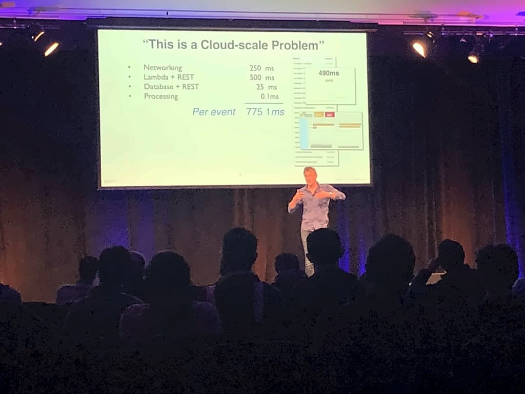 A speaker on a dark stage presenting a latency breakdown slide headed "This is a Cloud-scale Problem", totalling 775 milliseconds per event