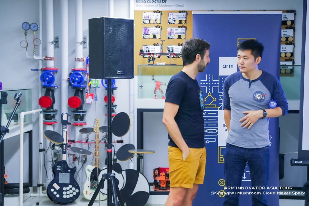 Two men talking in front of an Arm Innovator Program banner, beside a wall of maker-built instruments including a drum kit and a custom guitar