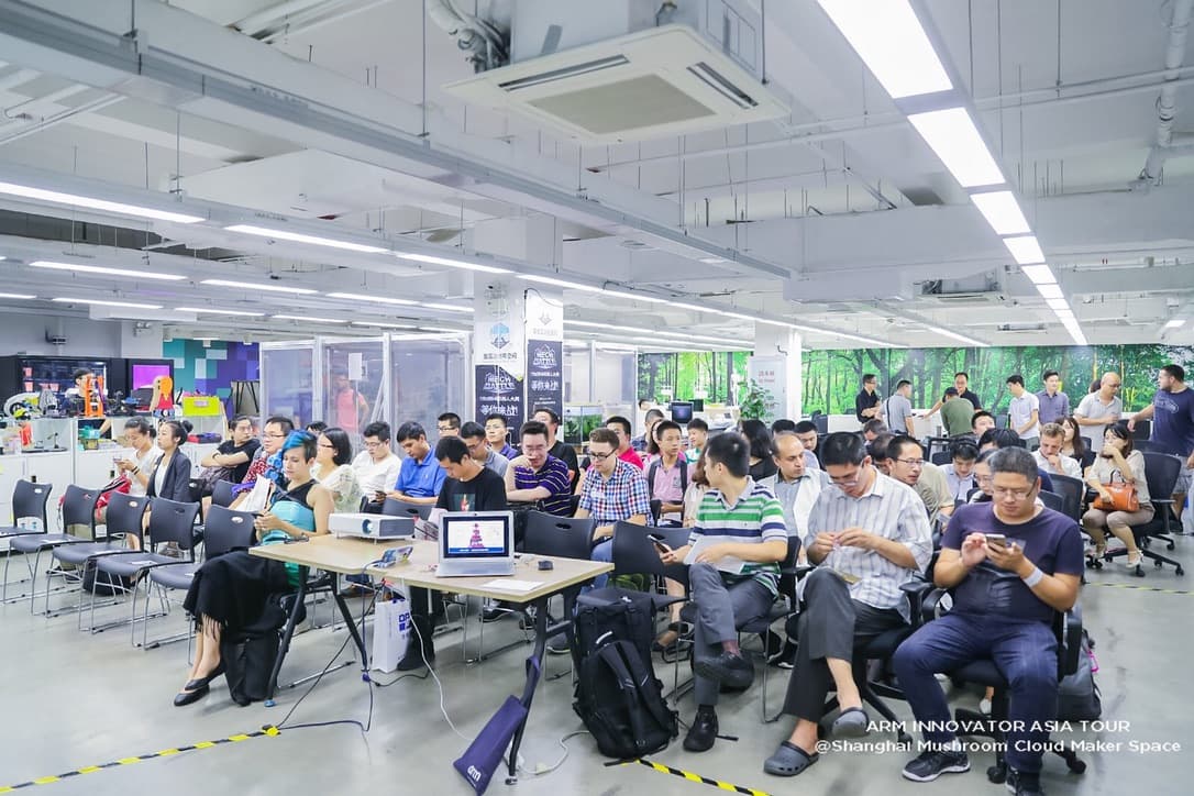 A seated audience of about sixty in a maker space, facing a table with a projector and a laptop