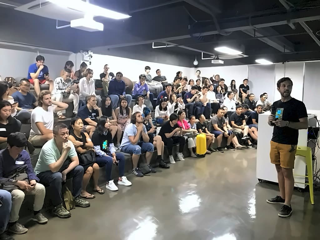 A speaker holding up a development board to a packed room seated on the floor and on sofas