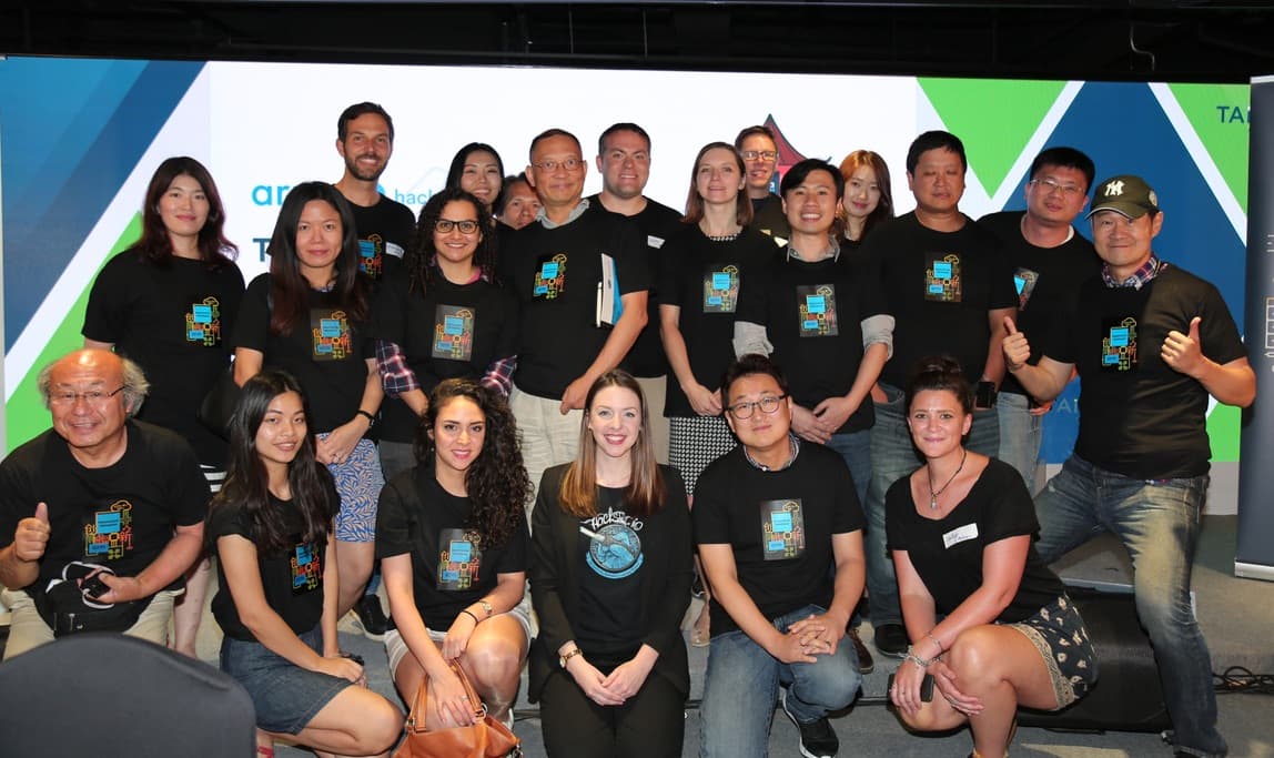 About twenty people in matching black tour T-shirts posing in front of an Arm and Hackster.io backdrop