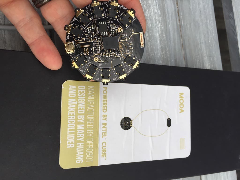 The MODA board beside its product card, reading powered by Intel Curie, designed by Mary Huang and MakerCollider, manufactured by DFRobot