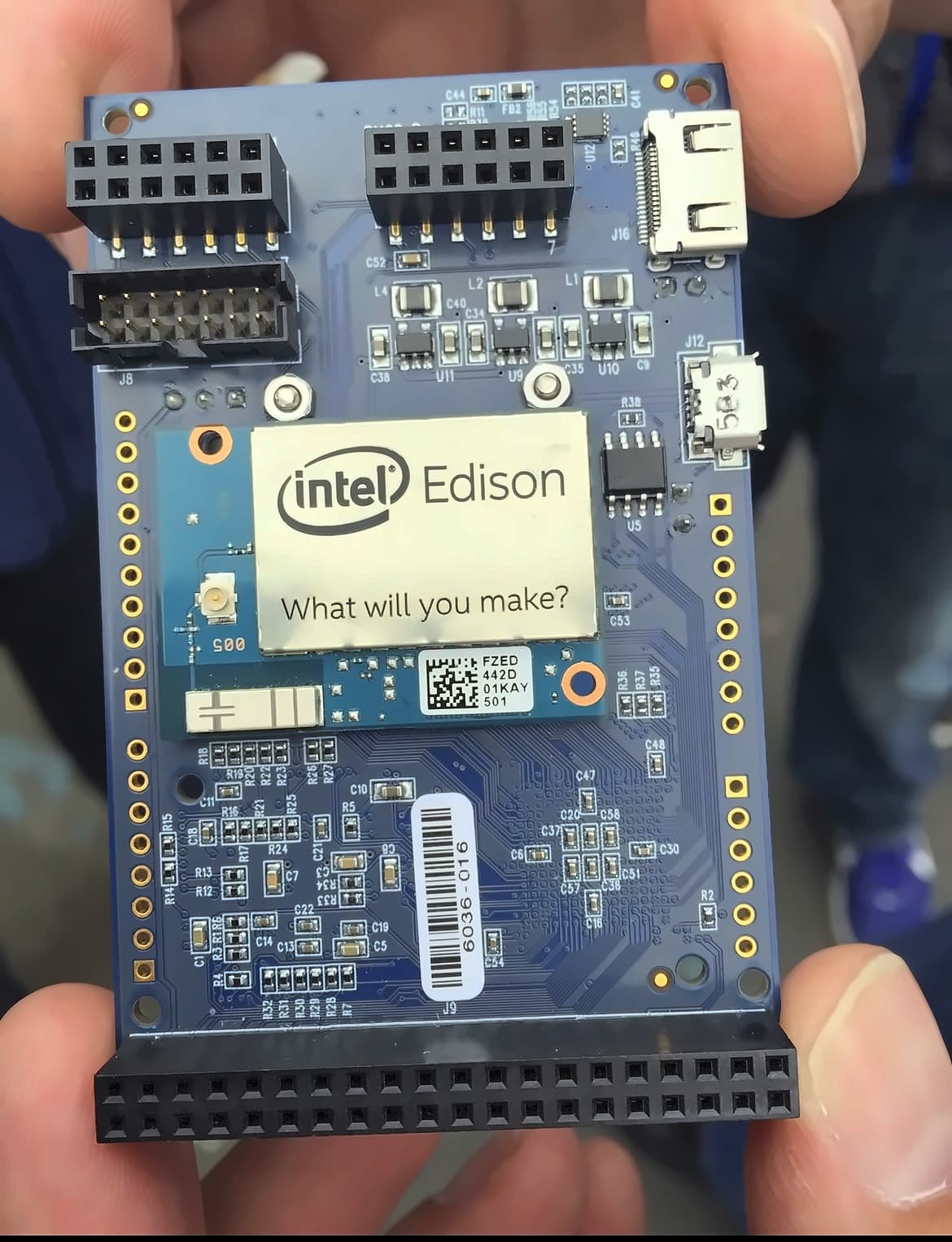 A hand holding an Edison on a blue breakout board with header pins, a micro SD slot and micro USB