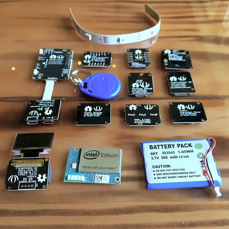 A bare Edison module laid out with a dozen Seeed Studio Xadow components — OLED screen, barometer, buzzer, vibration motor, NFC tag, LED strip and a lithium battery pack