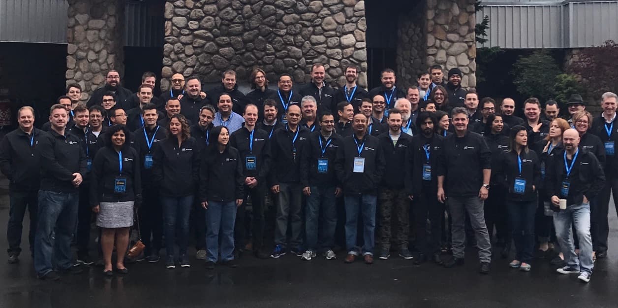 About seventy people in matching black jackets and blue lanyards in a group photo on wet pavement in front of a stone building