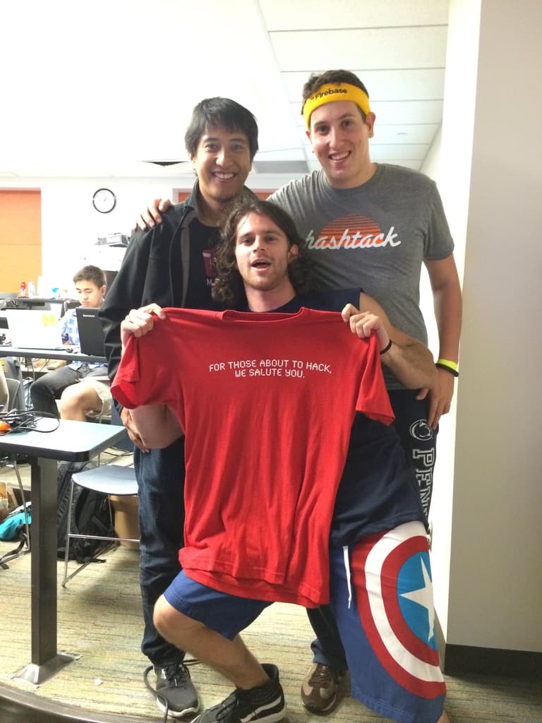 Three hackathon participants grinning at the camera, the one in front holding up a red t-shirt reading "for those about to hack, we salute you"