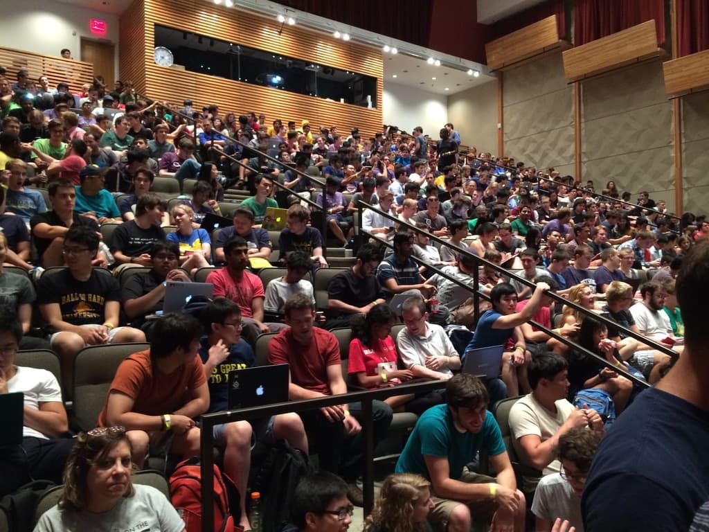 A university lecture hall filled to the back rows with several hundred hackathon participants, laptops open across every tier of seating