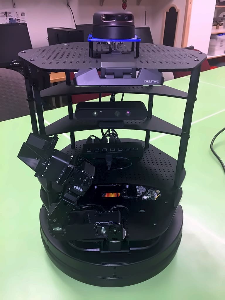 A multi-tier mobile robot on a lab bench, carrying a depth camera, a Creative sensor bar and a second depth camera across three decks above a circular drive base