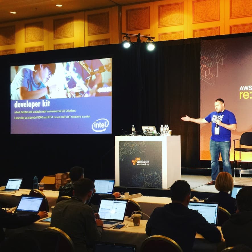 Rex presenting at AWS re:Invent in front of a screen reading "developer kit — a fast, flexible and scalable path to commercial IoT solutions" with the Intel logo, an AWS re:Invent banner behind him and rows of attendees with open laptops