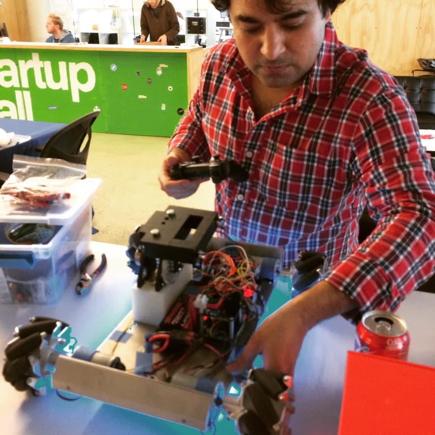 A developer wiring a wheeled robot chassis with mecanum wheels at a table, a Startup Hall sign behind him