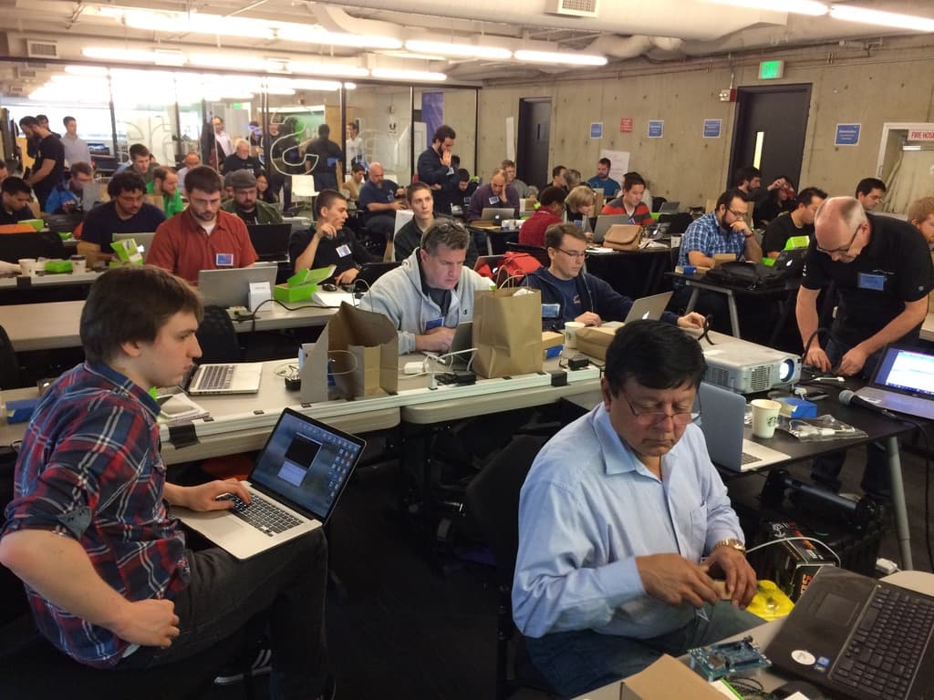 A long room of developers working at laptops on shared tables, several unpacking boards and kits from paper bags