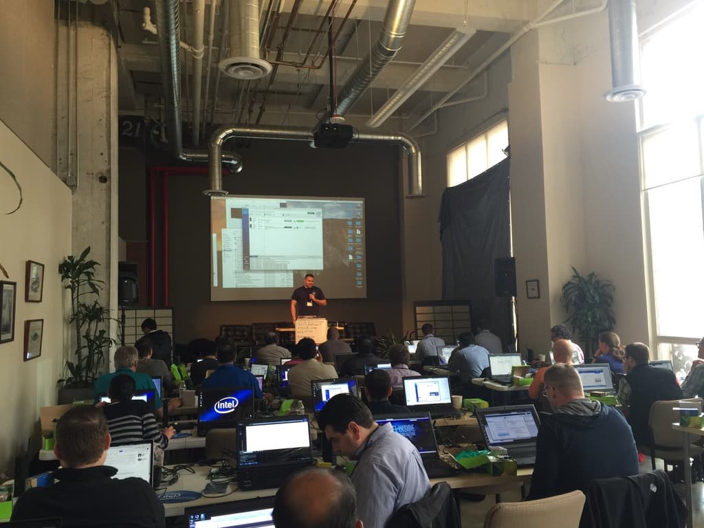A full workshop room with around thirty attendees at long tables, every laptop open and Intel-branded, green developer kit boxes beside them, Rex presenting at the front with a terminal projected behind him