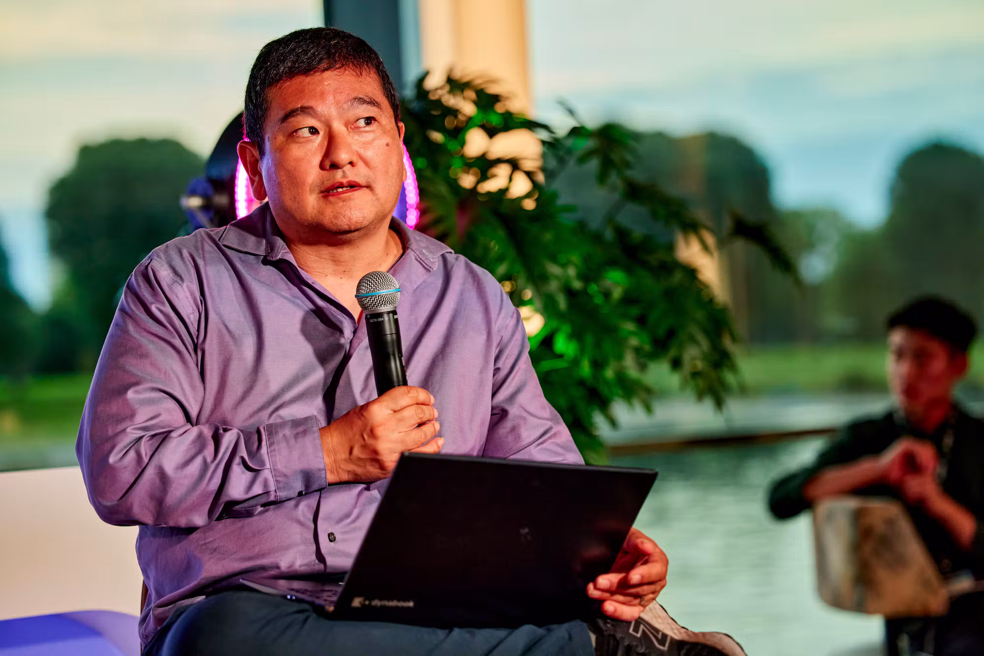 A speaker with a microphone and open laptop on stage, the Rhine visible through the window behind him